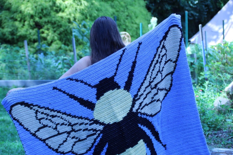 May include: A crocheted blanket featuring a large bee design. The bee is black, yellow, and white, with wings that are beige and black. The blanket is a light blue color.