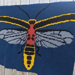 May include: A crocheted blanket featuring a large, colorful insect with yellow, black, red, and gray details. The insect is depicted on a dark blue background.
