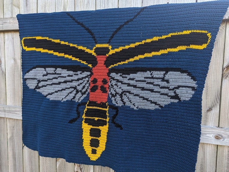 May include: A crocheted blanket featuring a large, colorful insect with yellow, black, red, and gray details. The insect is depicted on a dark blue background.