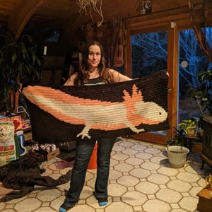 May include: A woman holds a crocheted blanket featuring a pink and white axolotl on a black background. The blanket is made with white and black yarn.