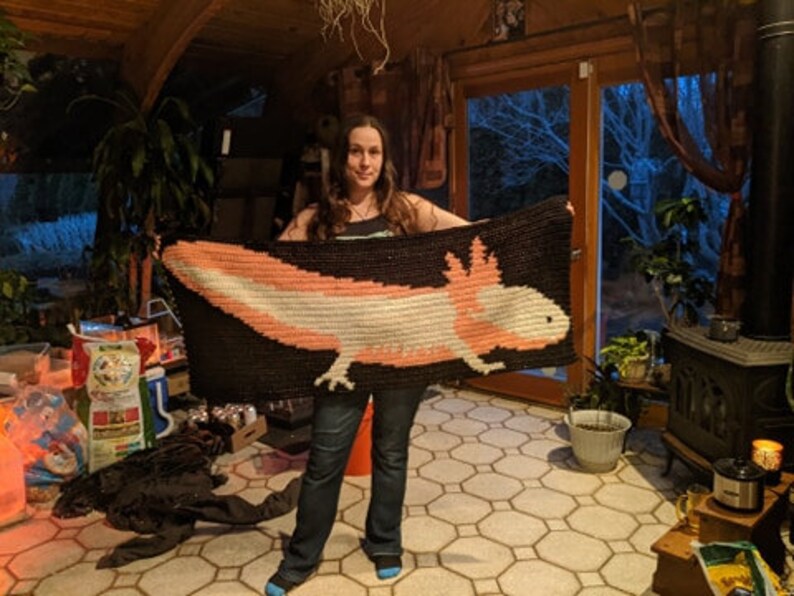 May include: A woman holds a crocheted blanket featuring a pink and white axolotl on a black background. The blanket is made with white and black yarn.