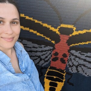 May include: A woman stands next to a crocheted wall hanging featuring a large, colorful moth with yellow, red, black, and gray details.