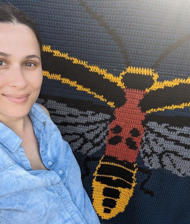 May include: A woman stands next to a crocheted wall hanging featuring a large, colorful moth with yellow, red, black, and gray details.