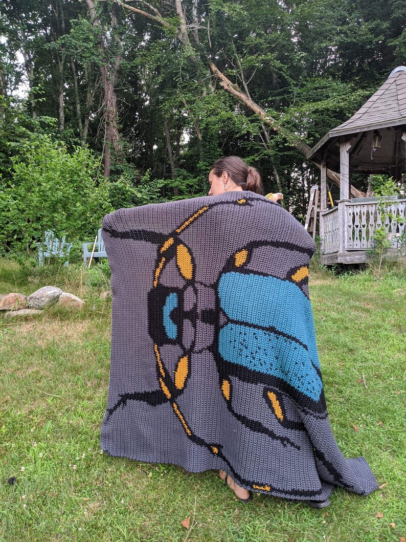 May include: A gray crocheted blanket with a large blue and yellow beetle design. The blanket is being held up by a person in a grassy area.