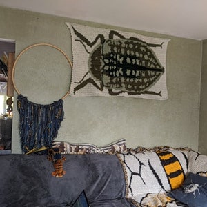 May include: A living room with a crocheted wall hanging of a beetle, a macrame dream catcher, and a crocheted throw blanket with a bee design on a couch.