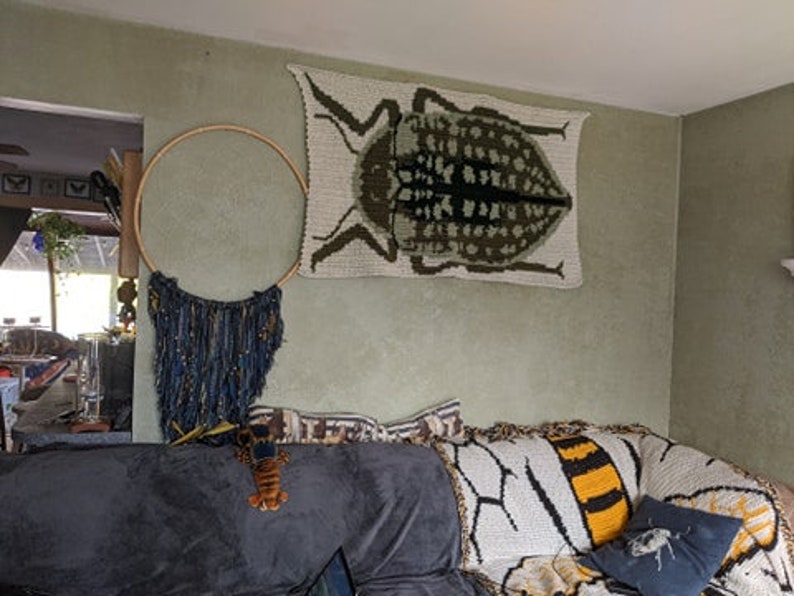 May include: A living room with a crocheted wall hanging of a beetle, a macrame dream catcher, and a crocheted throw blanket with a bee design on a couch.