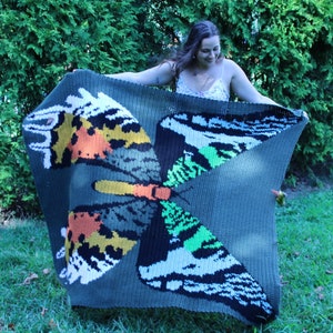 May include: A crocheted blanket featuring a large, colorful butterfly design. The butterfly has wings with shades of green, orange, yellow, black, and white. The blanket is a dark green color.