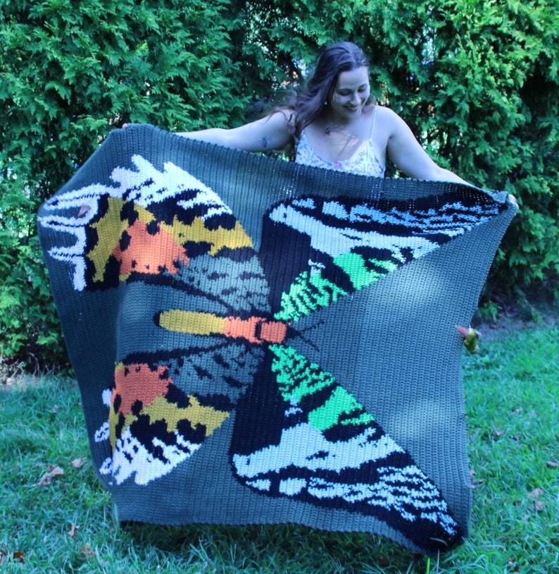 May include: A crocheted blanket featuring a large, colorful butterfly design. The butterfly has wings with shades of green, orange, yellow, black, and white. The blanket is a dark green color.