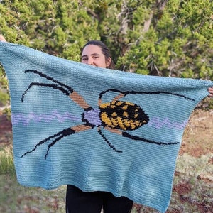 May include: A blue crocheted blanket with a yellow and black spider design. The blanket is being held by a person in front of a green background.