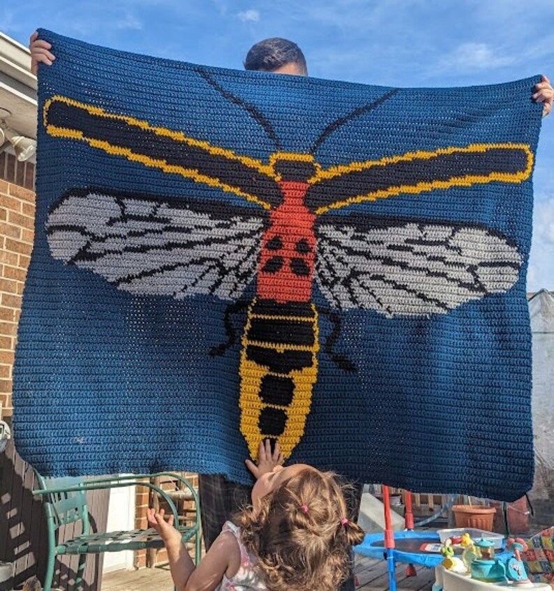 May include: A crocheted blanket featuring a large, colorful insect with yellow, red, black, and white details. The blanket is blue and has a crocheted texture.