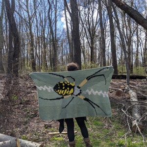 May include: A person is holding a crocheted blanket with a green background and a yellow and black spider design.
