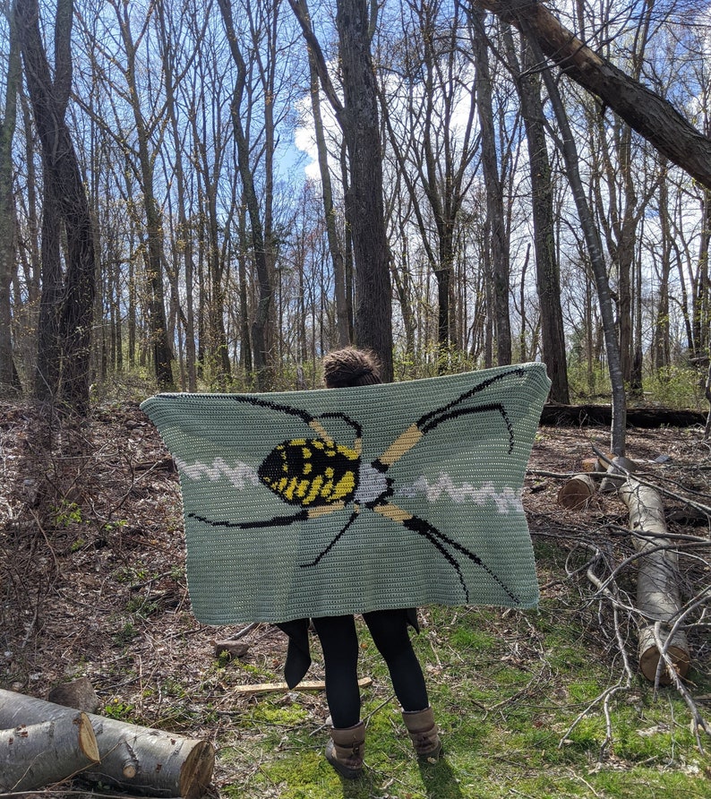 May include: A person is holding a crocheted blanket with a green background and a yellow and black spider design.