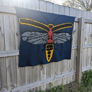 May include: A crocheted blanket featuring a large, colorful insect with yellow, red, and black accents. The insect is depicted on a dark blue background.
