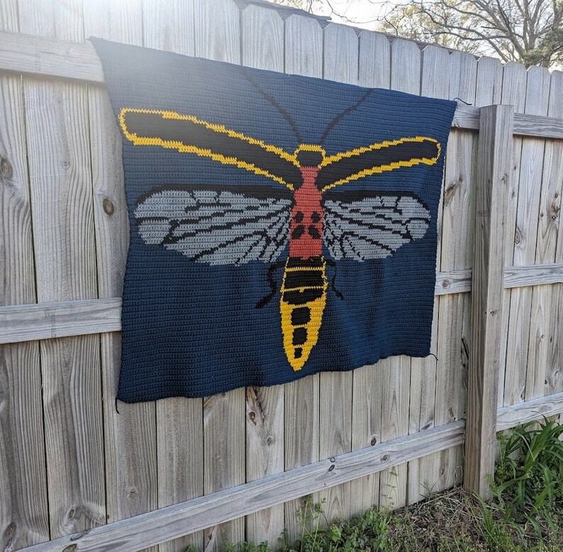 May include: A crocheted blanket featuring a large, colorful insect with yellow, red, and black accents. The insect is depicted on a dark blue background.