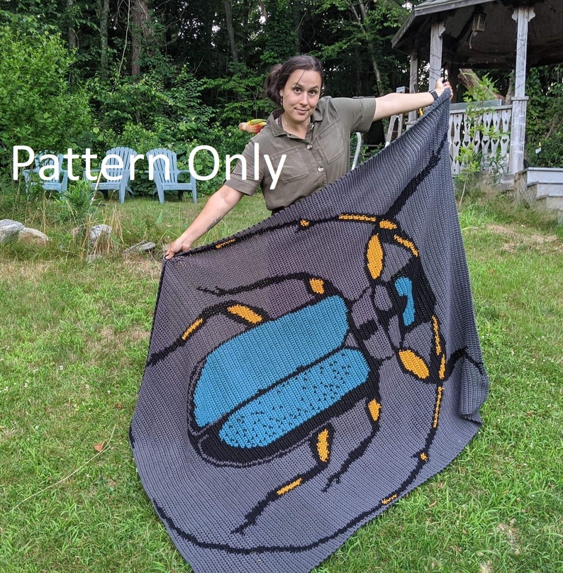 May include: A crocheted blanket featuring a large, detailed beetle design. The beetle is blue, black, and yellow, and is set against a gray background. The blanket is being held up by a person in a green shirt.