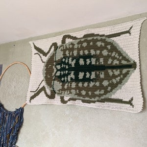 May include: A crocheted wall hanging featuring a large, detailed beetle in shades of brown, green, and white.