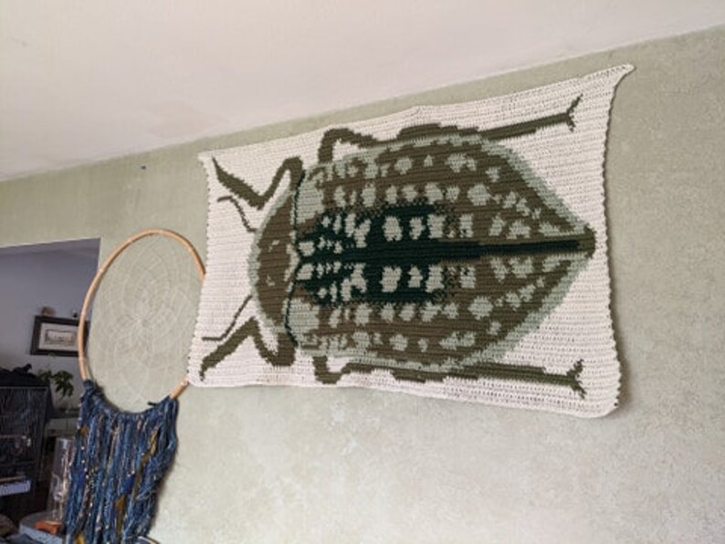 May include: A crocheted wall hanging featuring a large, detailed beetle in shades of brown, green, and white.