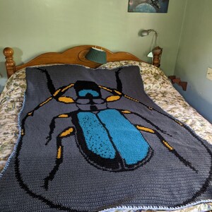 May include: A gray crocheted blanket with a large blue and yellow beetle design. The blanket is on a bed with a floral patterned bedspread.
