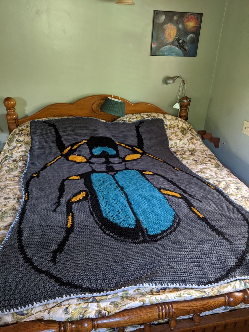 May include: A gray crocheted blanket with a large blue and yellow beetle design. The blanket is on a bed with a floral patterned bedspread.