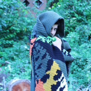 May include: A person wearing a hooded crocheted blanket with a colorful geometric pattern. The blanket is made of green, black, orange, yellow, and white yarn.
