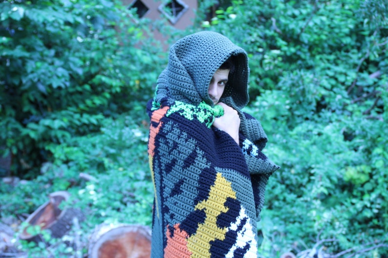 May include: A person wearing a hooded crocheted blanket with a colorful geometric pattern. The blanket is made of green, black, orange, yellow, and white yarn.