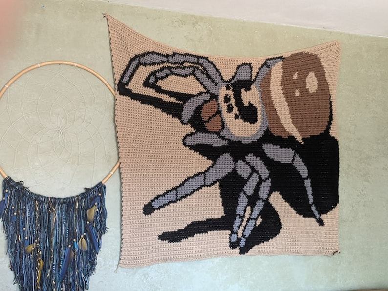 Op de afbeelding: Een gehaakte wandhanger met een grote spin in tinten bruin, zwart en grijs. De spin is afgebeeld met acht poten en een groot, rond lichaam.