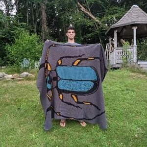 May include: A person holding a gray crocheted blanket with a large blue and yellow beetle design.