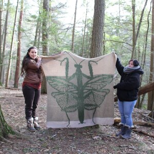 May include: Two people are holding up a large crocheted blanket featuring a green praying mantis. The blanket is beige with a green praying mantis design. The people are standing in a wooded area.