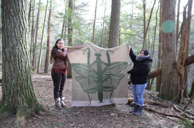 May include: Two people are holding up a large crocheted blanket featuring a green praying mantis. The blanket is beige with a green praying mantis design. The people are standing in a wooded area.