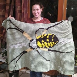 May include: A crocheted blanket featuring a large yellow and black spider with a green background. The blanket is being held by a person.