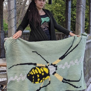 May include: A green crocheted blanket with a black and yellow wasp design. The blanket is being held by a person with long brown hair.