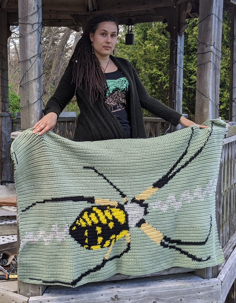 May include: A green crocheted blanket with a black and yellow wasp design. The blanket is being held by a person with long brown hair.
