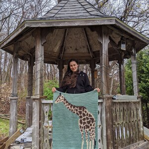 May include: A woman stands in a wooden gazebo holding a crocheted blanket featuring a giraffe design. The blanket is light blue with a brown and white giraffe.