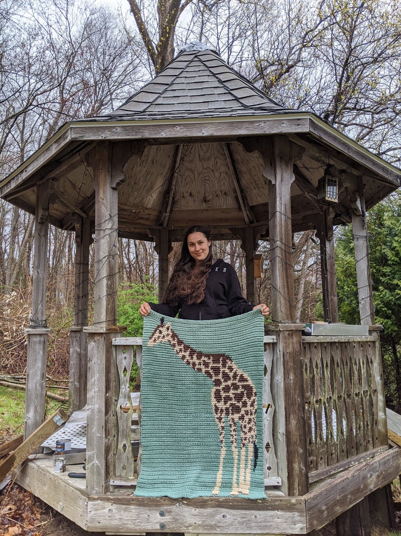 May include: A woman stands in a wooden gazebo holding a crocheted blanket featuring a giraffe design. The blanket is light blue with a brown and white giraffe.