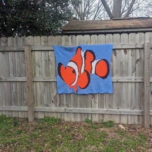 May include: A crocheted wall hanging featuring a cartoon clownfish with orange, white, and black details on a blue background.