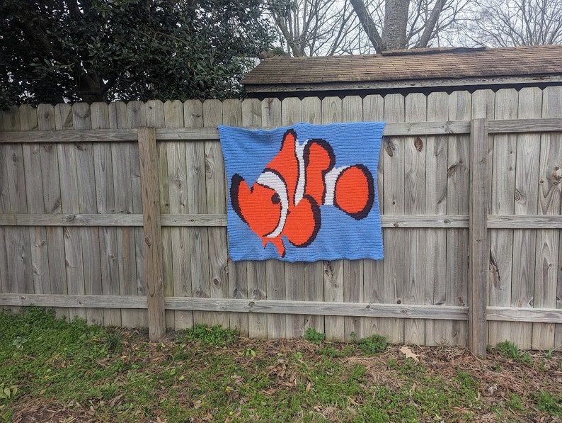May include: A crocheted wall hanging featuring a cartoon clownfish with orange, white, and black details on a blue background.