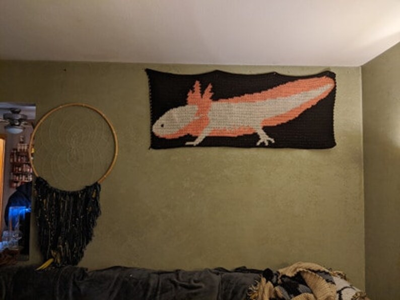 May include: A crocheted wall hanging featuring a pink and white axolotl on a black background.