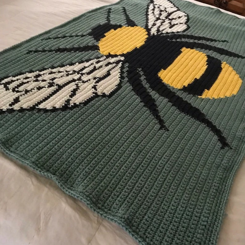 May include: A crocheted blanket featuring a large bee design. The bee is yellow, black, and white, and is set against a green background.