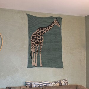 May include: A crocheted wall hanging featuring a giraffe in brown and black against a teal green background.
