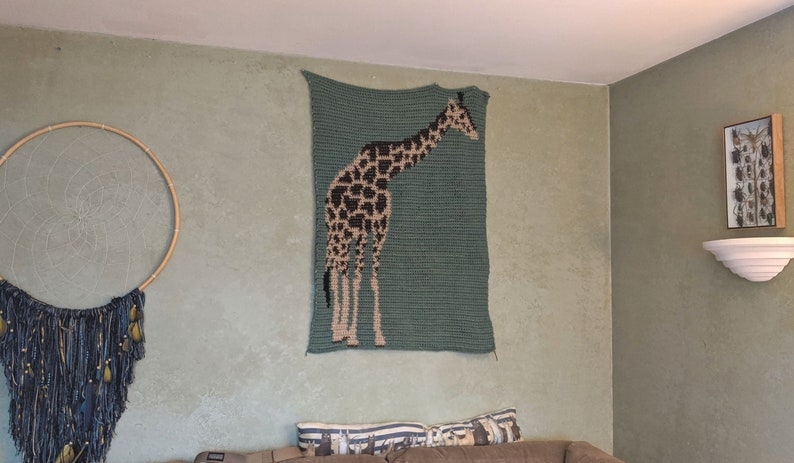 May include: A crocheted wall hanging featuring a giraffe in brown and black against a teal green background.