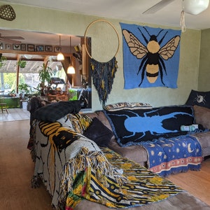 May include: A living room with a couch covered in blankets with bee and beetle designs. The couch is in front of a wall with a blue tapestry featuring a bee and a macrame hanging on the wall.