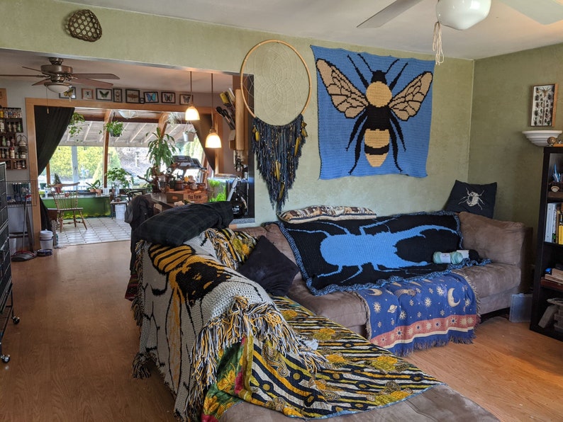May include: A living room with a couch covered in blankets with bee and beetle designs. The couch is in front of a wall with a blue tapestry featuring a bee and a macrame hanging on the wall.