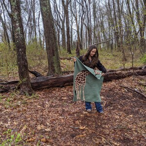 May include: A woman in a woodland setting holds up a green crocheted blanket featuring a giraffe design. The blanket is folded in half and she is holding it in front of her.
