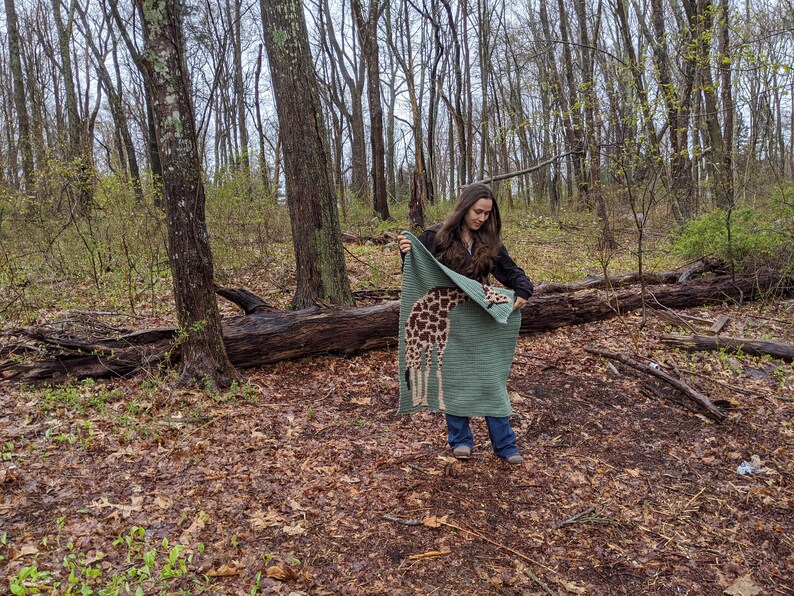 May include: A woman in a woodland setting holds up a green crocheted blanket featuring a giraffe design. The blanket is folded in half and she is holding it in front of her.