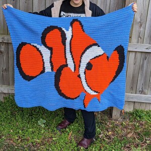 May include: A crocheted blanket featuring a large orange, white, and black clownfish design on a blue background.