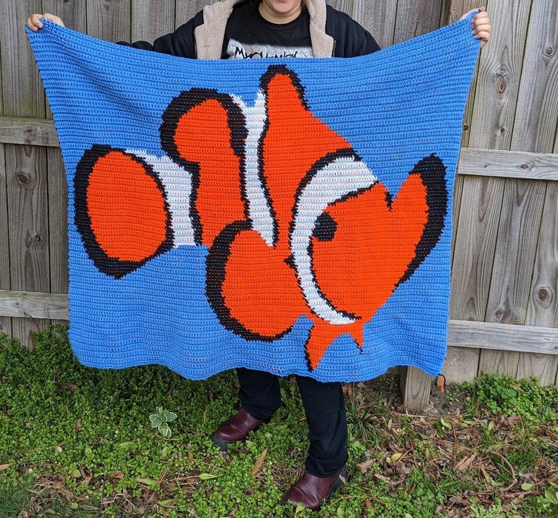 May include: A crocheted blanket featuring a large orange, white, and black clownfish design on a blue background.