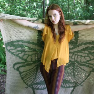 May include: A woman holds a crocheted blanket with a green butterfly design. The blanket is beige and green with a crocheted texture. The woman is wearing a yellow top and striped pants.
