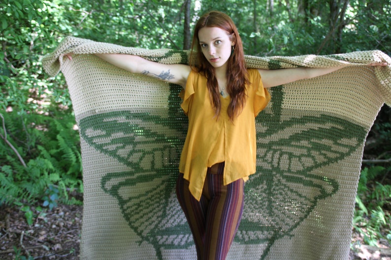 May include: A woman holds a crocheted blanket with a green butterfly design. The blanket is beige and green with a crocheted texture. The woman is wearing a yellow top and striped pants.