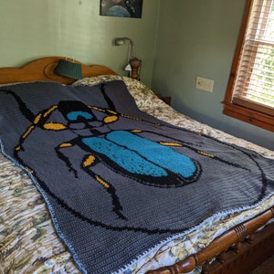 May include: A gray crocheted blanket with a large blue and black beetle design. The beetle has yellow accents and is centered on the blanket.