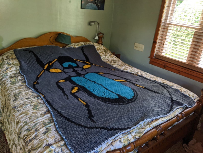May include: A gray crocheted blanket with a large blue and black beetle design. The beetle has yellow accents and is centered on the blanket.